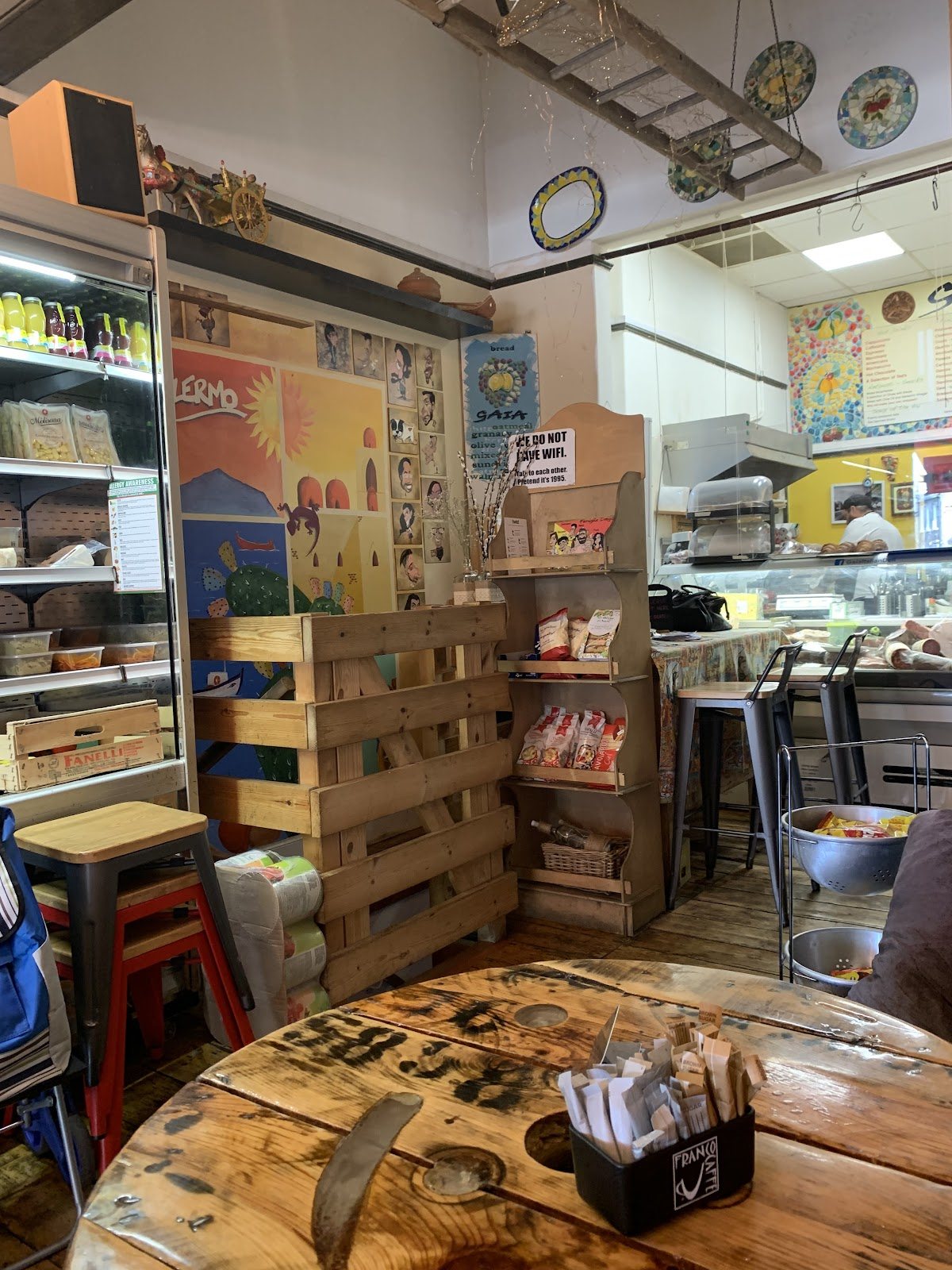 Inside Gaia: a spool table in front, wooden pallet shelving, red stools, a drinks fridge, painted ceramic plates on the wall and the deli counter at the back.