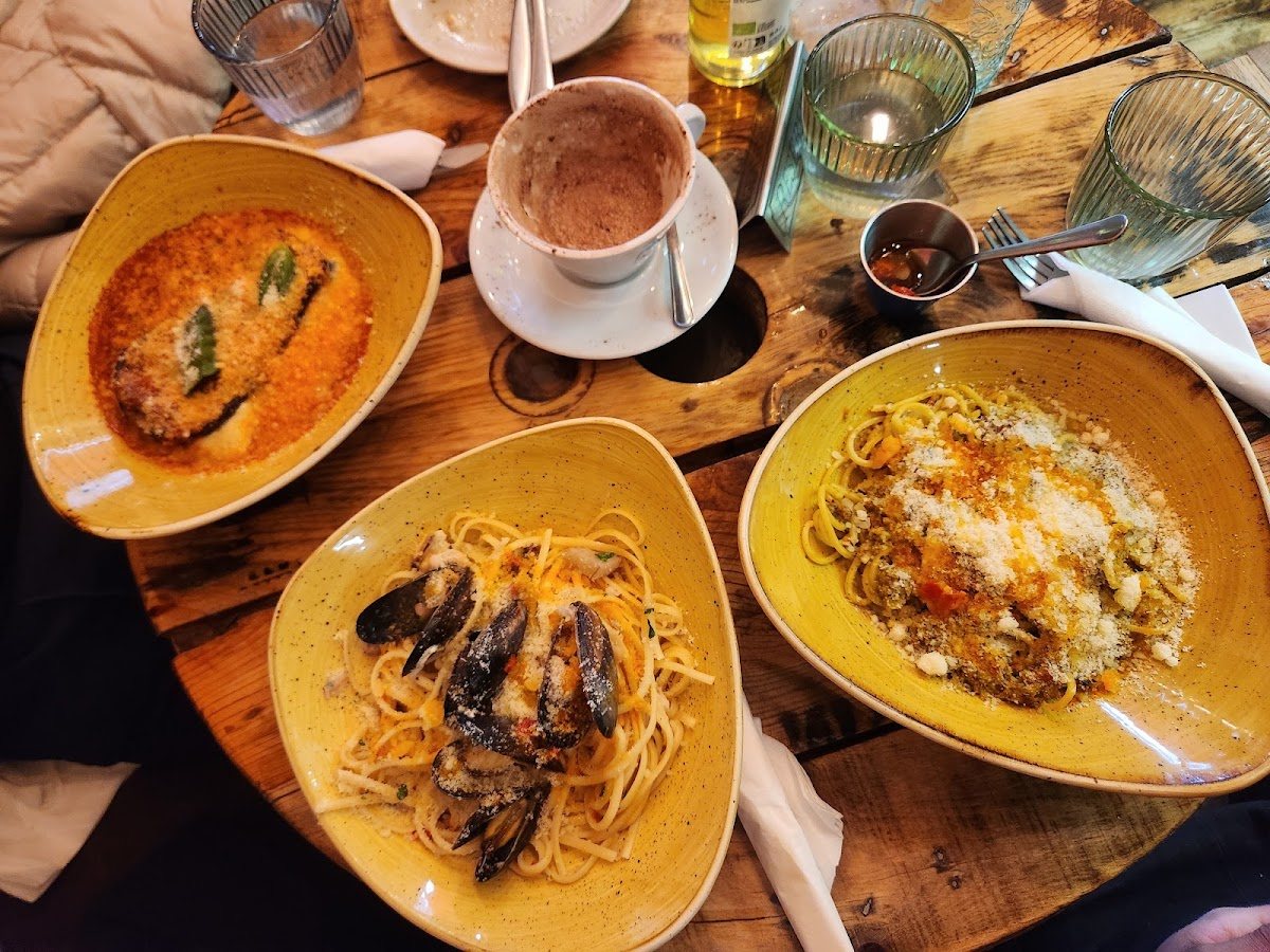 Three yellow bowls on a wooden table: spaghetti with mussels, pasta in tomato with toasted crumbs, and aubergine in tomato sauce.