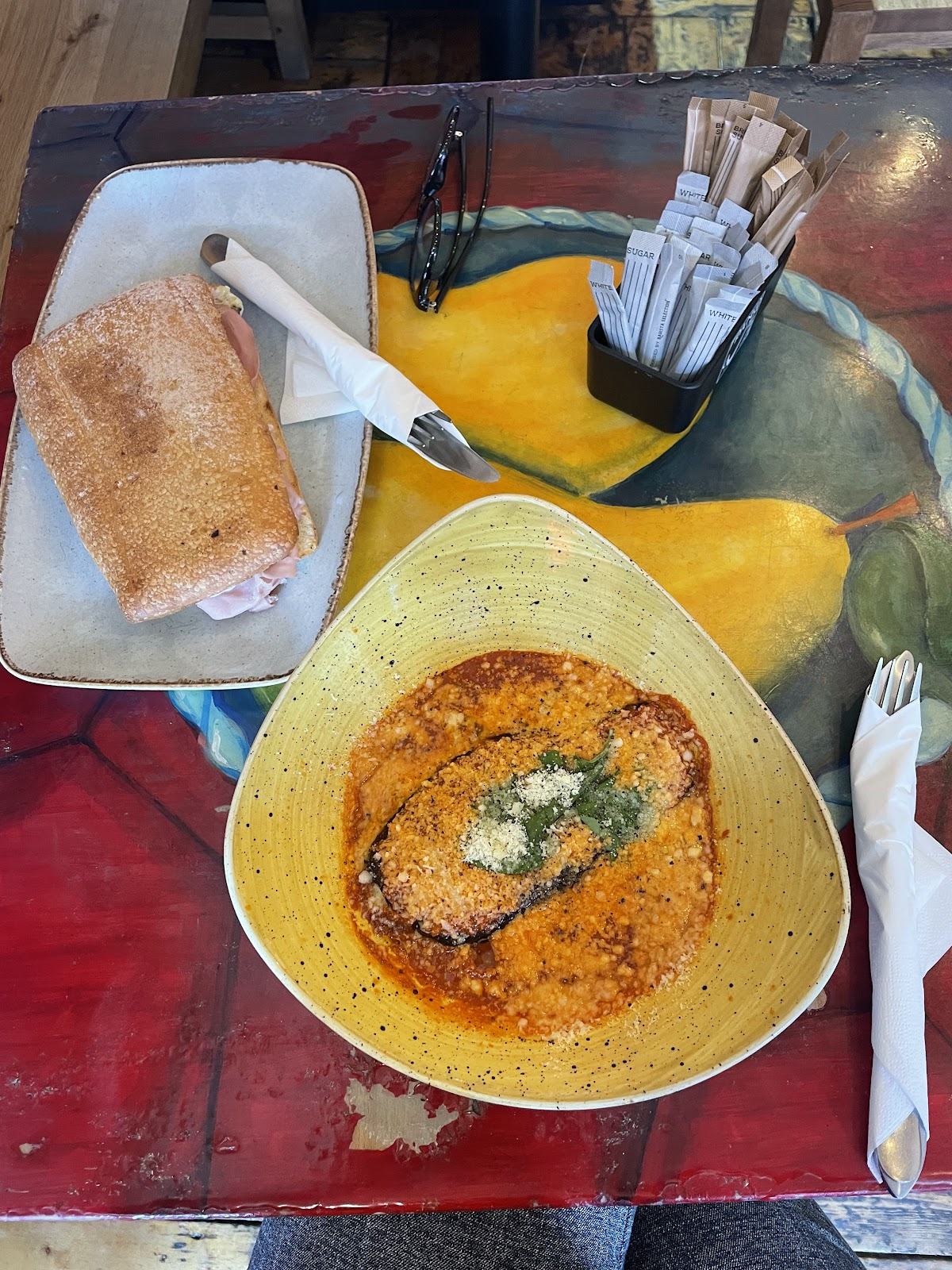 Aubergine baked in tomato sauce with grated cheese and herbs in a yellow bowl, beside a toasted panino, on a painted table.